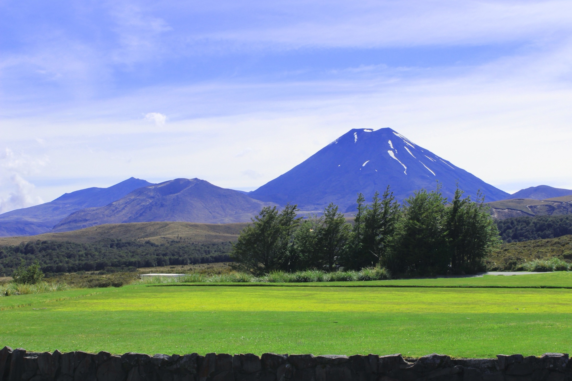 A snow-dusted volcanic mountain rising behind green fields and native trees along Te Araroa
